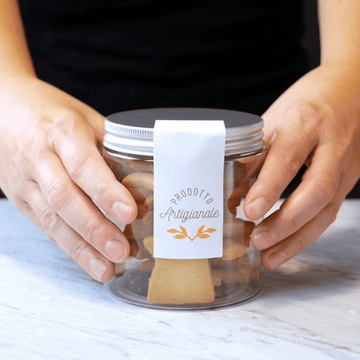 Vasetto trasparente con tappo in alluminio pieno di biscotti ed adesivo prodotto artigianale retto tra due mani